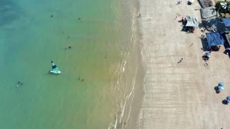 Bird's-eye-aerial-shot-of-the-tropical-Bessa-beach-with-turquoise-clear-water-in-the-capital-city-of-Joao-Pessoa,-Paraiba,-Brazil-with-people-enjoying-the-ocean,-sand,-and-a-sail-boat-on-a-summer-day