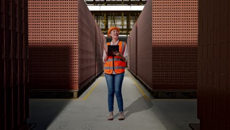 full body of asian female engineer with safety helmet looking at the tablet in her hand and looking around while standing with red brick packed in stacks are stored