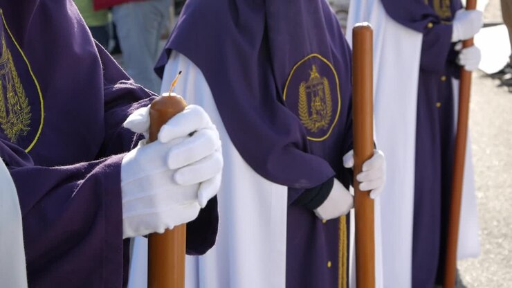 Primo piano di tre penitenti in fila, vestiti con tuniche tradizionali bianche e viola, pronti a marciare.