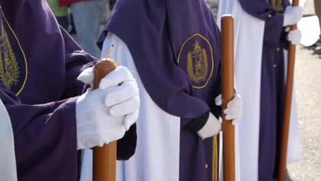 Primer-Plano-De-Tres-Penitentes-En-Formación-En-Línea-Vestidos-Con-Túnicas-Tradicionales-Blancas-Y-Moradas,-Listos-Para-Marchar.