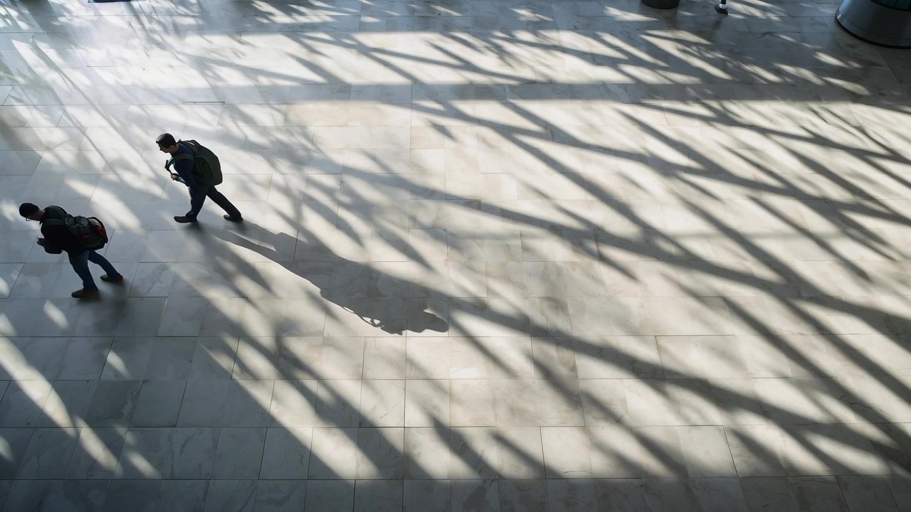 Entering upper-left, two men commuting in hall under shadows, backpacks, checking phone, copy space