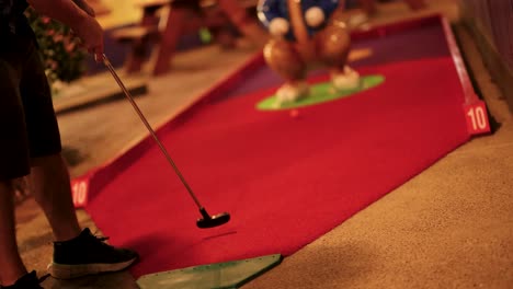 a person putts on a red carpeted mini-golf course at night, illuminated by warm, ambient lighting