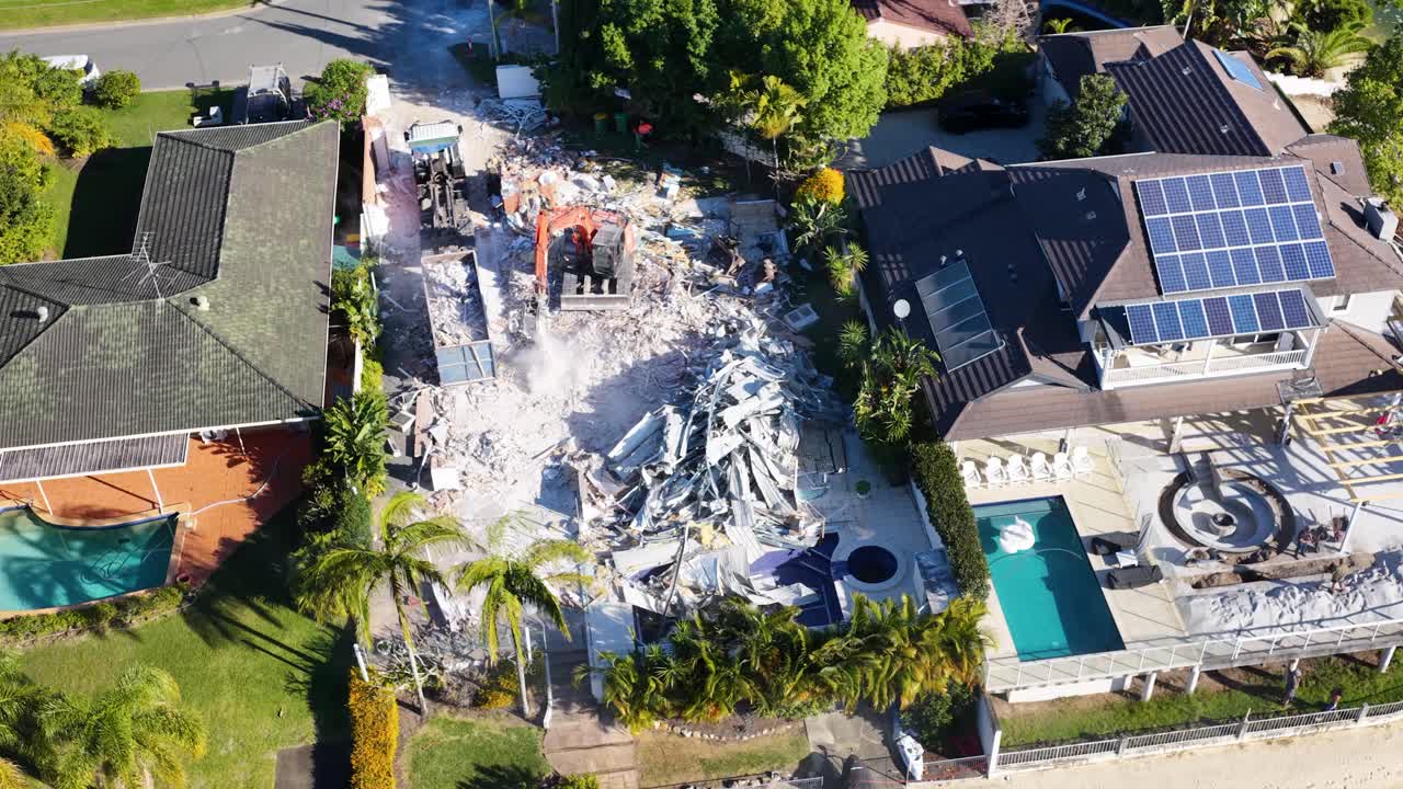 Drone footage captures a waterfront house being demolished by an excavator in bright daylight, with debris and dust visible near neighboring homes and a pool