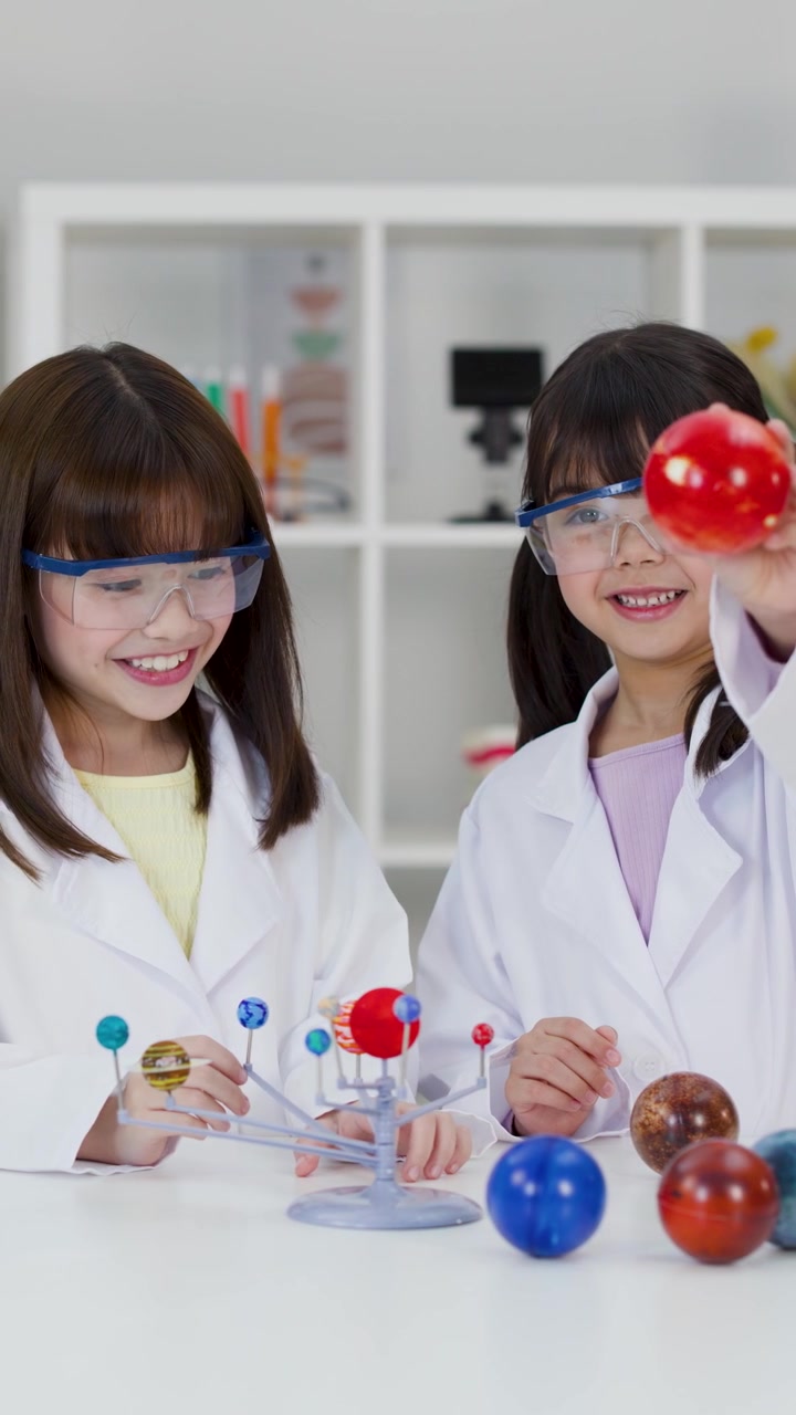 Dua siswa muda mengenakan kacamata pengaman memeriksa model planet selama pelajaran sains di kelas.