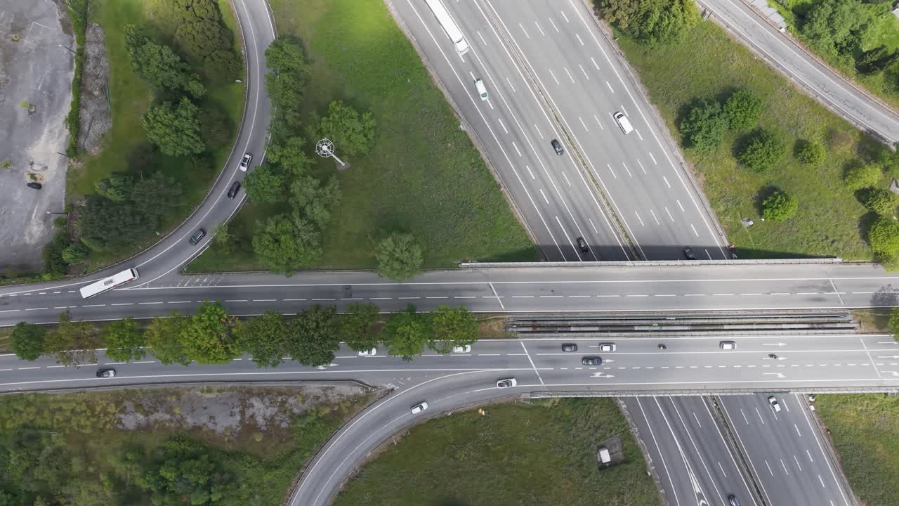 Dynamic aerial shot of cars and trucks on a multi-lane highway interchange. The drone follows traffic flow, showing motion, roads, and green areas.