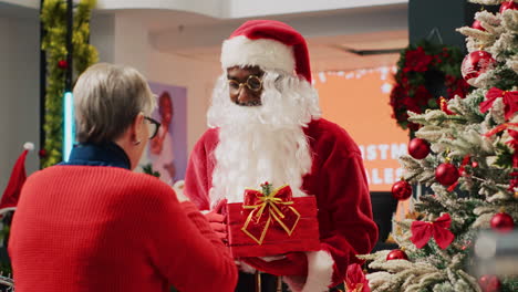 Älteres-Ehepaar-In-Einem-Weihnachtlich-Geschmückten-Bekleidungsgeschäft,-Das-Einem-Als-Weihnachtsmann-Verkleideten-Arbeiter,-Der-Ein-Geschenk-Hält-Und-Als-Sammelbox-Dient,-Ein-Tombola-Los-überreicht.-Mitarbeiter-Organisiert-Weihnachtswerbeveranstaltung