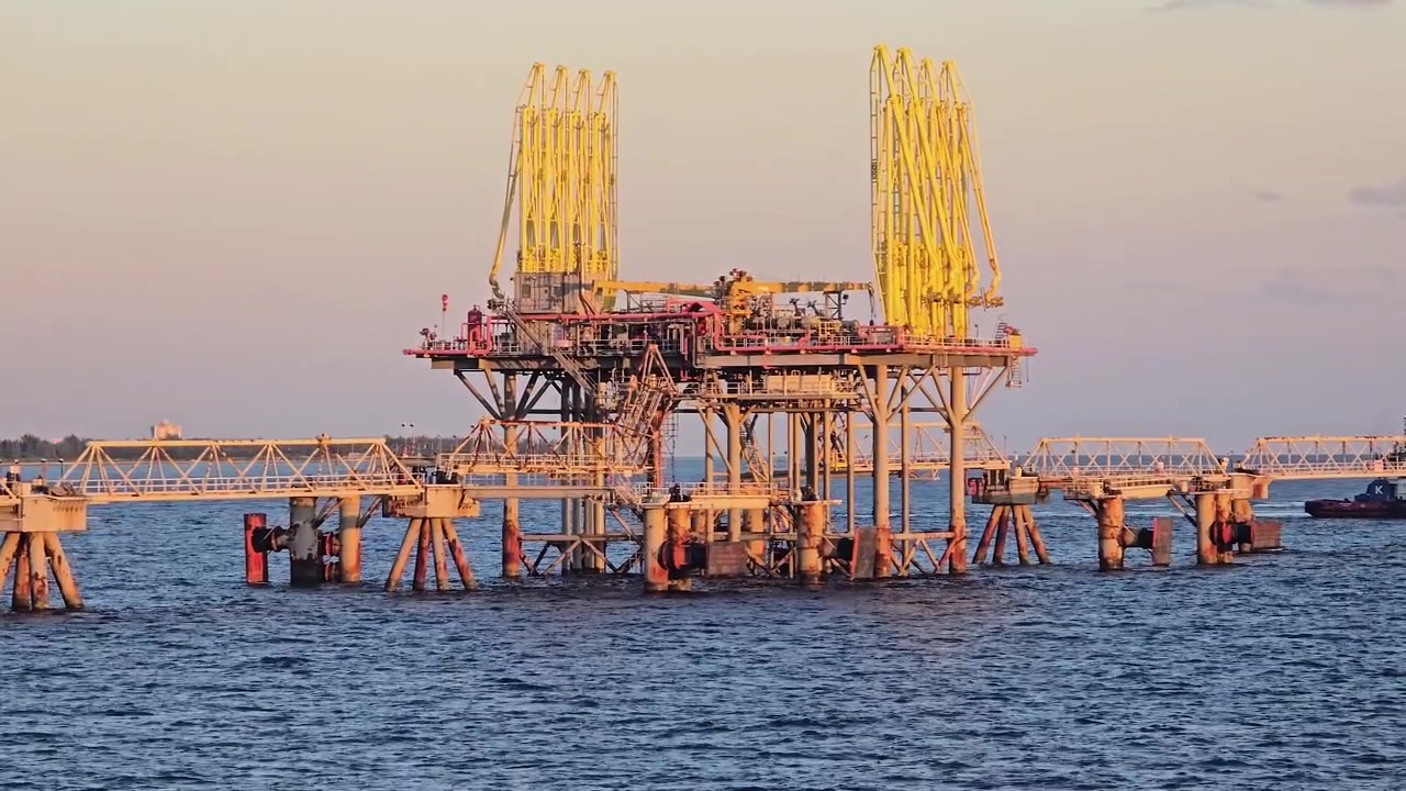 Close shot of offshore industrial oil terminal platform stands in the blue ocean during sunset. Large yellow loading arms connect to a pier structure while a small boat navigates nearby