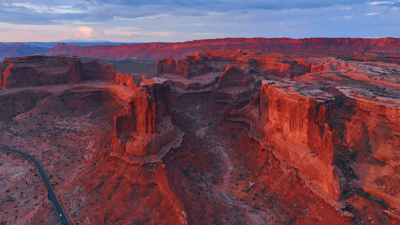 Red walls of the majestic canyons are lit with setting sun. Cars ride by the desert in the Arches National Park, Utah, USA. Aerial view