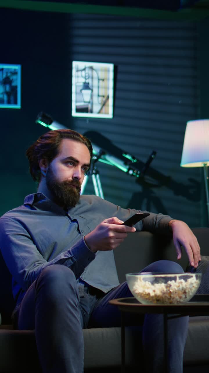Video verticale. Uomo sul divano che sfoglia film su un servizio di streaming usando il telecomando.