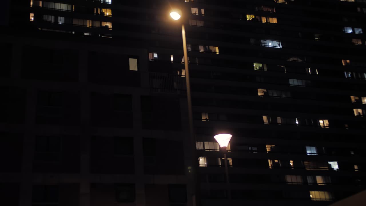 Moody urban shot from a moving car at night, capturing a towering apartment building with scattered glowing windows