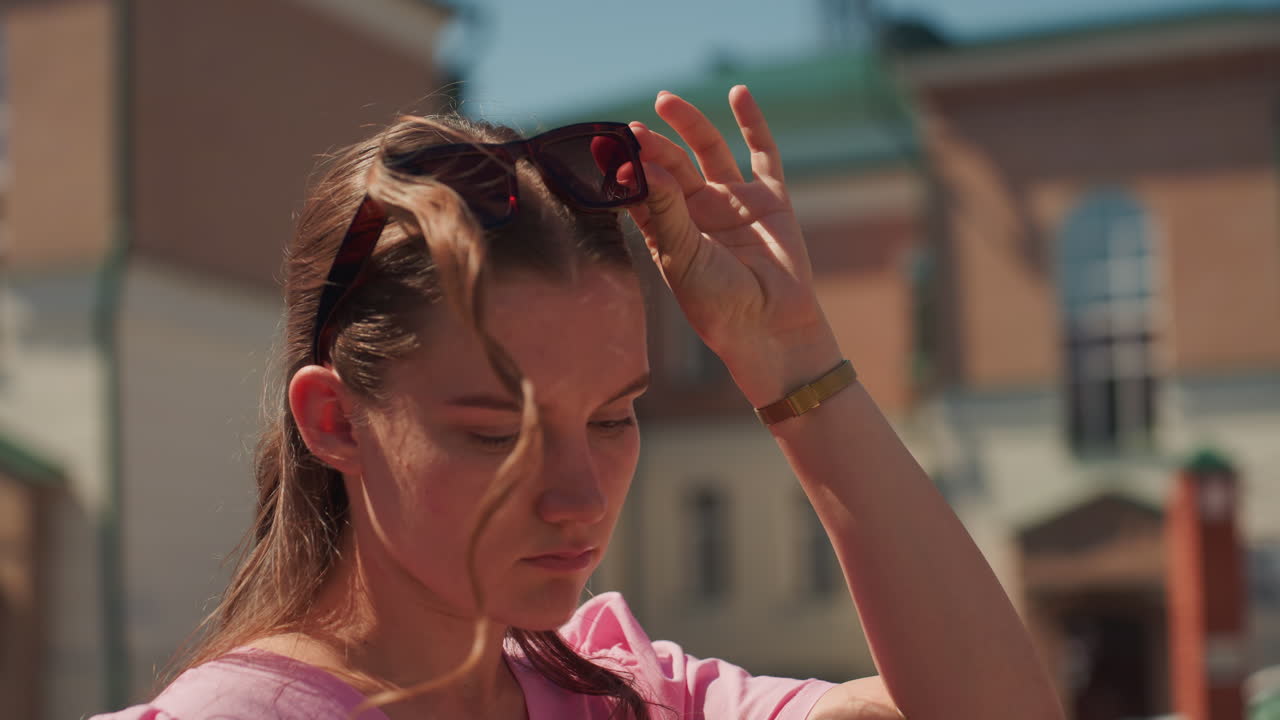 Giovane donna legge messaggi sul telefono in una strada soleggiata, occhiali da sole sulla testa, facciata in mattoni e auto parcheggiata sullo sfondo, primo piano che cattura l'espressione pensierosa e l'ottimizzazione del colore cinematografica, tranquillo contesto urbano