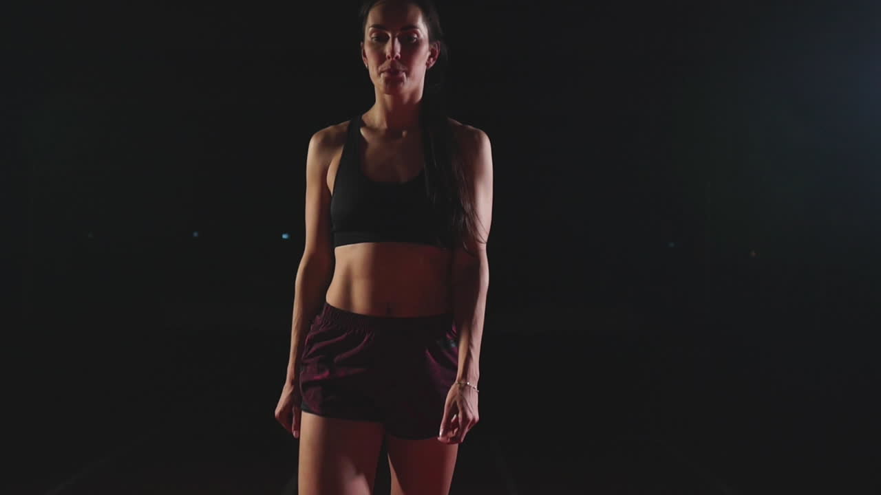 검은 쇼트와 운동화에 티셔츠를 입은 운동선수 여성이 스포츠 단지 트랙의 달리기 패드에 있고 느린 움직임으로 달리고 있습니다.