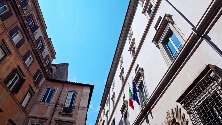 La telecamera si inclina verso l'alto tra vecchi condomini e un edificio civico bianco con bandiere italiane contro un cielo azzurro brillante.
