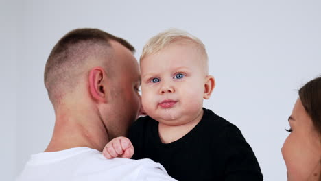 man holds a cute blond baby boy on his shoulder. lovely caucasian family close up. white backdrop.