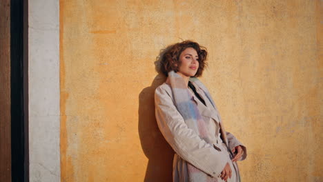 stylish woman leaning textured vintage wall in sunlight wearing coat closeup.