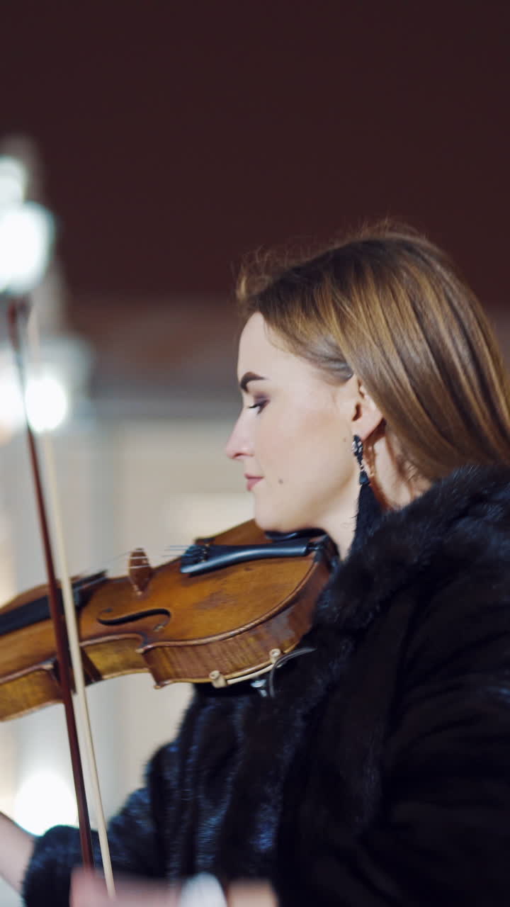A romantic girl in warm clothes is playing a violin in the street in the square on the background of urban architecture in the evening. Blurred background. Vertical video