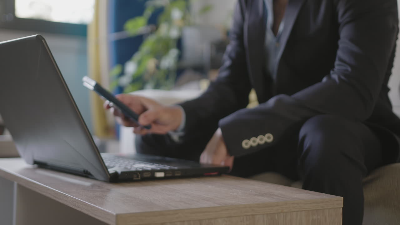 giovane uomo d'affari adulto che lavora da casa nel suo salotto usando un portatile, distratto dal lavoro con un telefono nuova e-mail pop-up, indossa il suo vestito prima di partire per il suo volo