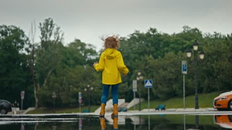 una adolescente feliz con una chaqueta amarilla y botas de goma naranja salta en un charco en el parque después de la lluvia