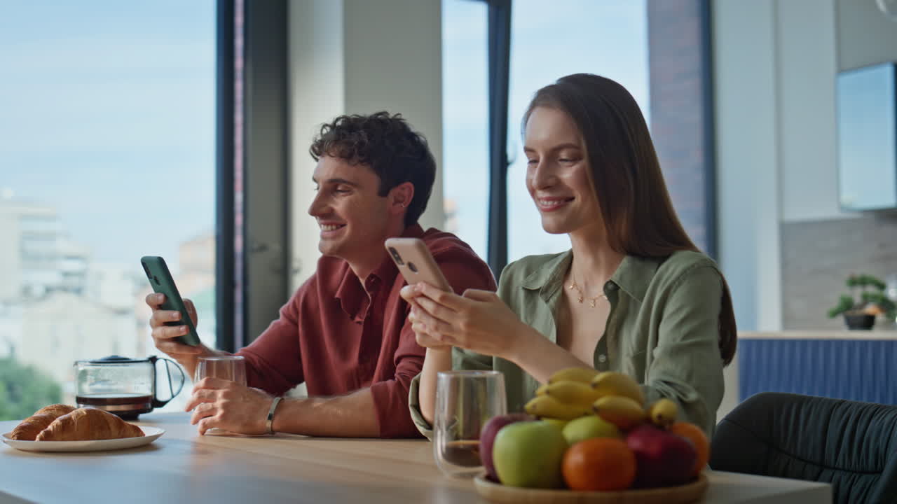 Young pair browsing smartphones sitting breakfast table together closeup