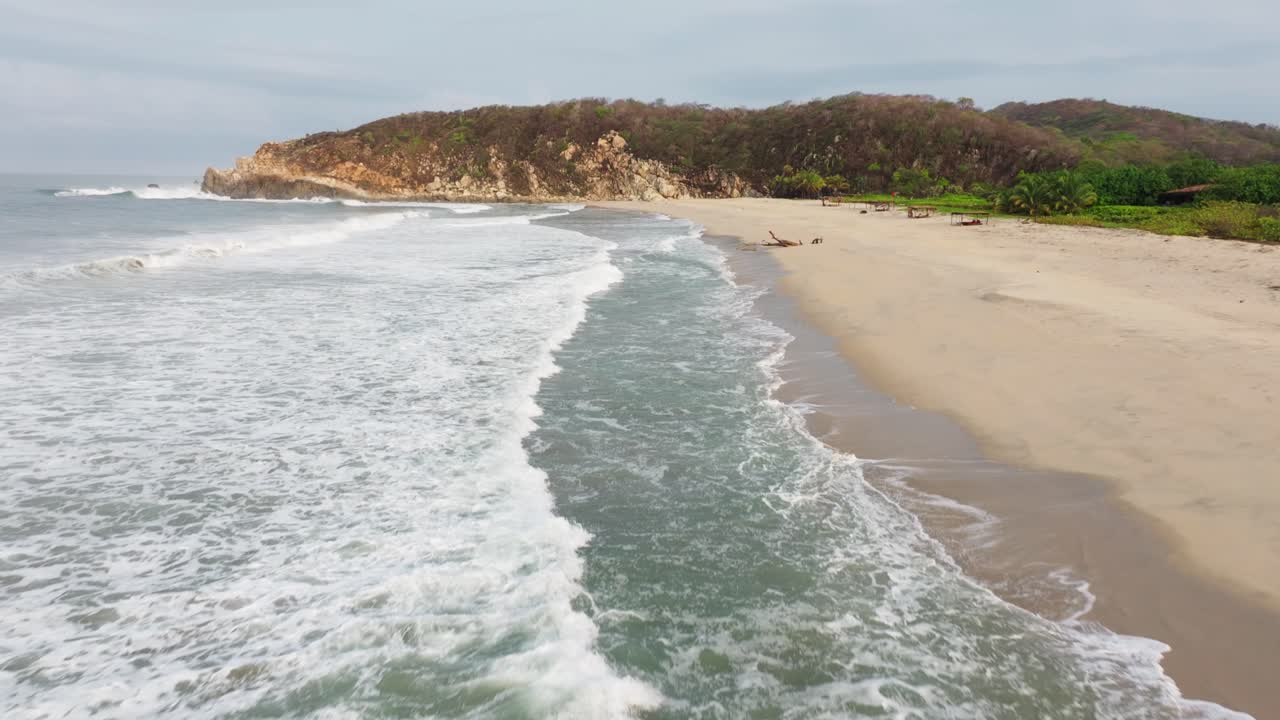 el dron se inclina hacia arriba mientras las olas del océano se estrellan y rodan sobre la playa de arena blanca con madera a la deriva y acantilados en el horizonte.