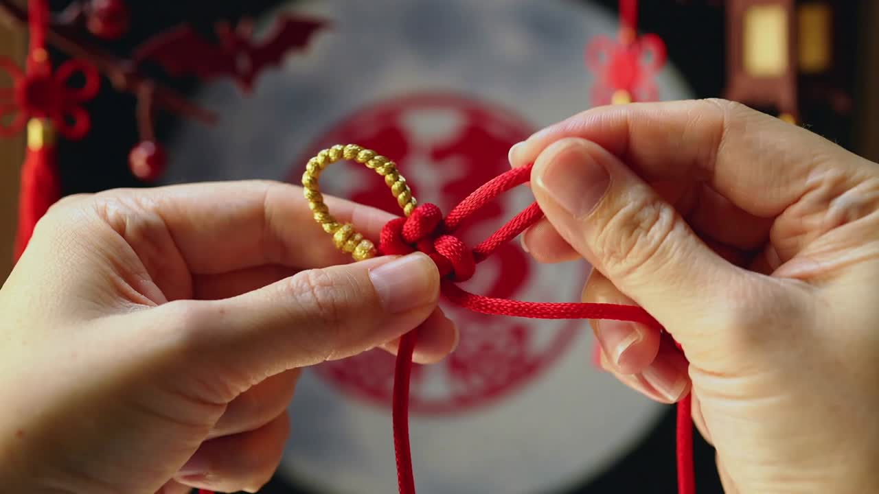 Mãos segurando um cordão vermelho e um aro dourado, formando um nó festivo em uma mesa de artesanato com franjas ao fundo