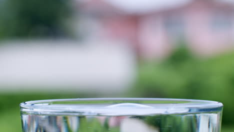 Wassertropfen-Treffen-Auf-Die-Oberfläche-Von-Kristallklarem-Wasser-In-Einem-Transparenten-Glasbecher-Auf-Einem-Verschwommenen-Hintergrund