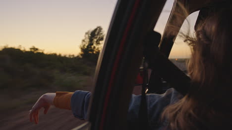 woman in car holding hand out window feeling wind blowing through fingers driving in countryside on road trip travelling for summer vacation enjoying freedom on the road at sunrise