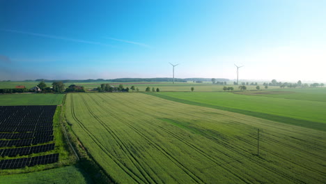 Toma-Panorámica-Aérea-Que-Muestra-Campos-Agrícolas-Con-Paneles-Solares-Instalados-Y-Molinos-De-Viento-Para-Energía-Verde-Limpia