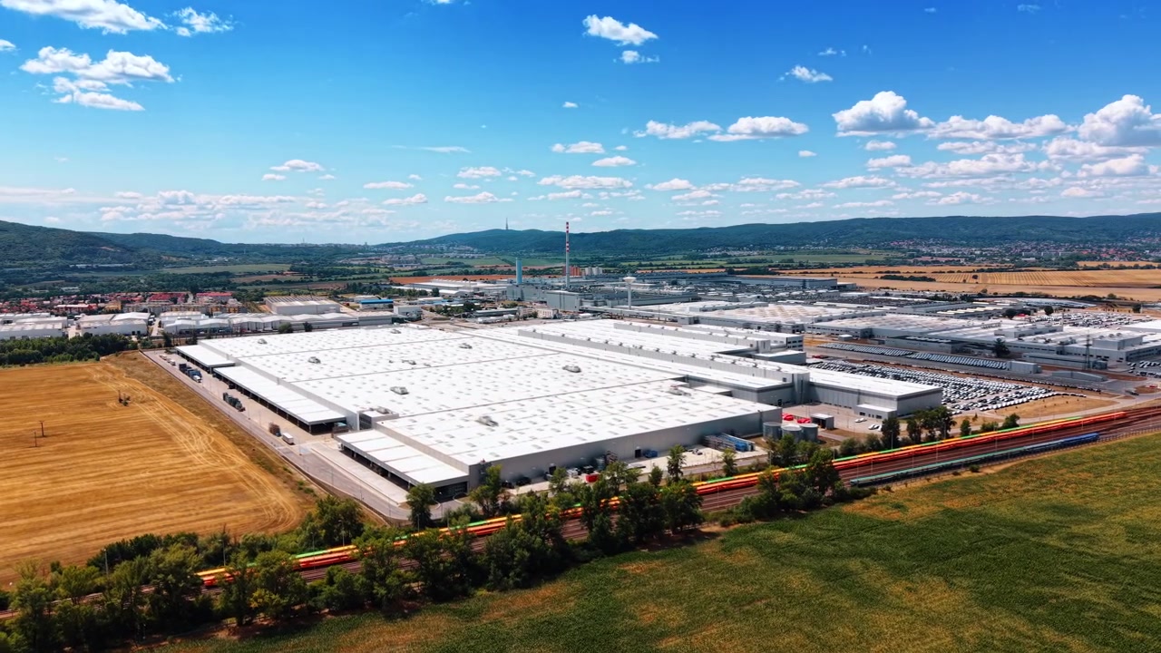 Grande stabilimento industriale circondato da terreni agricoli. Tetti industriali bianchi e vagoni ferroviari colorati si estendono tra colture verdi, campi raccolti e colline boscose.