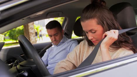 a man and a woman in a car. the woman is putting on her seat belt. the man is behind the wheel.