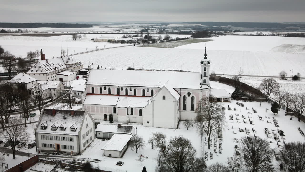 4K-drone flyver baglæns og viser den sneklædte Elchingen kirke, landsbyhuse, kirkegård og de store marker. Et roligt tilbagetræk giver et fredfyldt vinterlandskab – perfekt til dokumentarfilm, reklamer og visuelle elementer til turisme