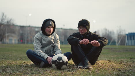 two boys are seated on a grassy field, engaged in a discussion, the boy wearing glasses is holding a stick, attempting to cut it, while the other boy in a gray top has a soccer ball in front of him