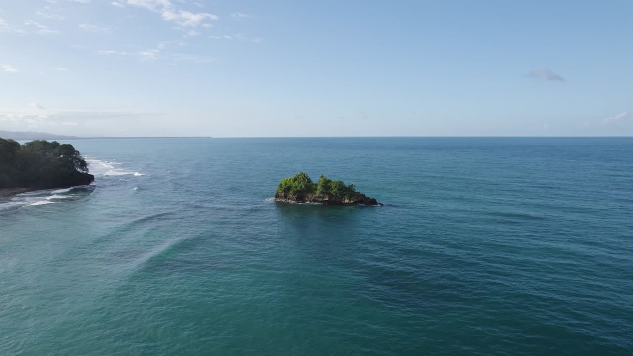 Aerial view of a small rock island with lush vegetation off the coast of Punta Cocles, Costa Rica, surrounded by the deep blue sea under a clear sky.