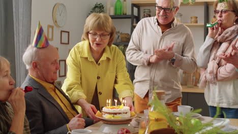 happy senior man blowing candles on birthday cake on dinner party