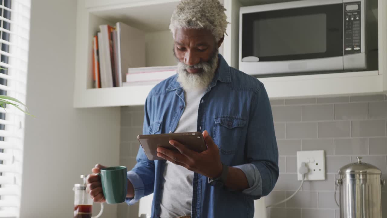 Video of happy african american senior man drinking coffee and using tablet