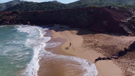 Drohnenansicht-Einer-Person,-Die-Mit-Ihrem-Hund-Am-Strand-Mit-Bergen-In-Spanien-Entlang-Geht