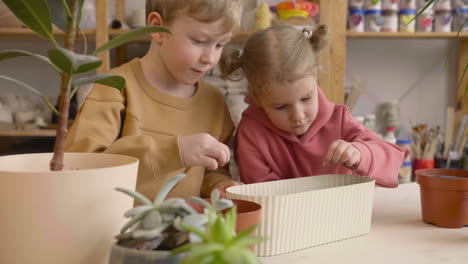 little blonde girl and blond kid preparing the soil in a pot sitting at a table where is plants in a craft workshop 3