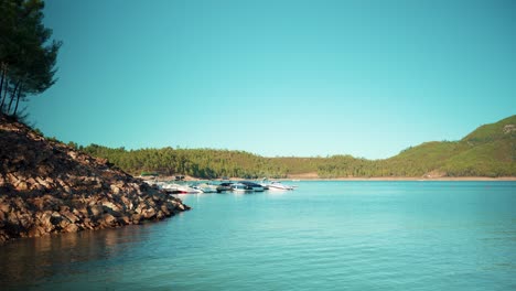 Lake-pier-with-parked-sport-boats-at-marina-with-mountain-pine-trees-under-blue-sky-at-sunshine-wide-shot-4K