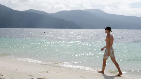 Ultra-slow-motion-shot-of-young-caucasian-man-with-cap-and-swimwear-walking-on-beach-on-sunny-day-in-Thailand