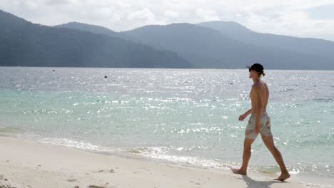 ultra slow motion shot of young caucasian man with cap and swimwear walking on beach on sunny day in thailand