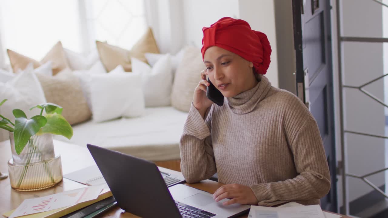 Video af smilende kvinde af blandet herkomst med hijab, der arbejder på bærbar computer og taler i smartphone derhjemme.