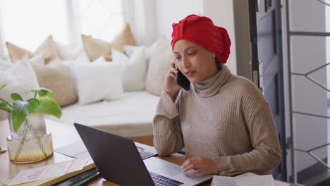 Video-Einer-Lächelnden-Gemischtrassigen-Frau-Im-Hijab-Am-Schreibtisch,-Die-Zu-Hause-Am-Laptop-Arbeitet-Und-Mit-Dem-Smartphone-Spricht