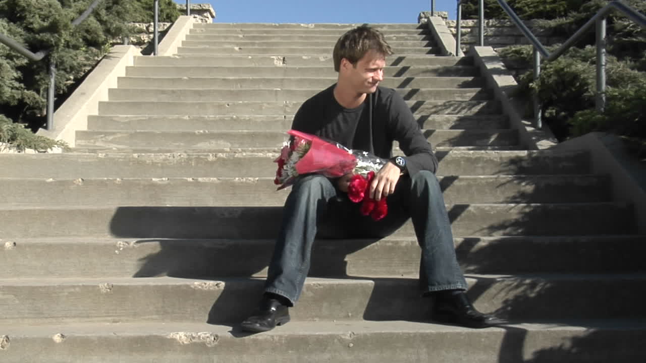 un hombre espera en las escaleras con flores y un oso de peluche