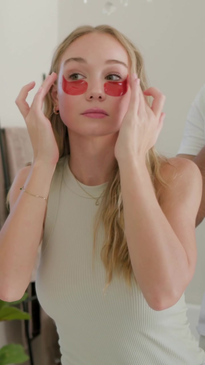 Vertical video: Picking up red patch, woman applying eye patches at vanity in tank top for skincare