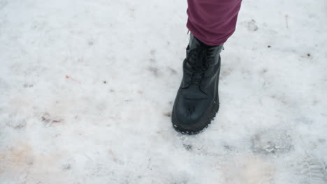 top-down perspective of individual in black boots playfully jumping on snow-covered ground with scattered dry foliage, winter setting highlights movement, footwear texture, and icy surface