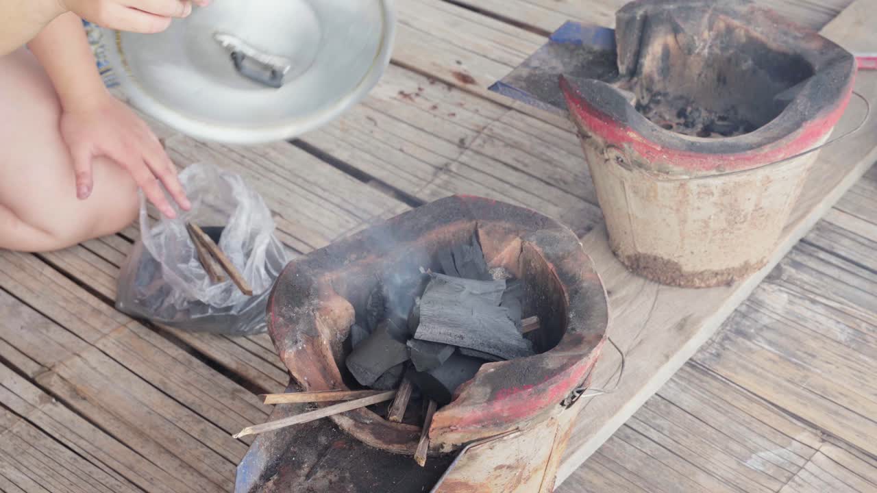 un primer plano de 4k de una estufa tradicional tailandesa de carbón con alguien usando una tapa de sartén para ventilar los carbones para un mejor calor con brasas fumando, creando una auténtica atmósfera de cocina tailandesa al aire libre en una cabaña de madera