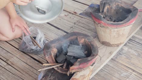 un primer plano de 4k de una estufa tradicional tailandesa de carbón con alguien usando una tapa de sartén para ventilar los carbones para un mejor calor con brasas fumando, creando una auténtica atmósfera de cocina tailandesa al aire libre en una cabaña de madera