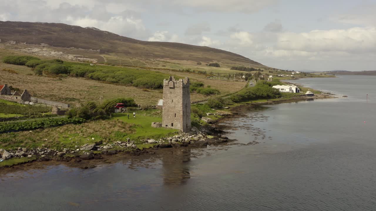 Aerial orbit around stone tower house reflecting in calm water below, cloudy day