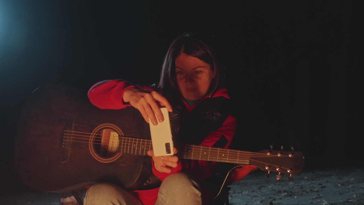 Natcamping med guitar ved bålet. Kvinde sidder med akustisk guitar på skødet og tager en smartphone-selfie ved siden af et varmt bål; glødende gløder og gnister oplyser et eftertænksomt ansigt, rød jakke og denim er synlige.