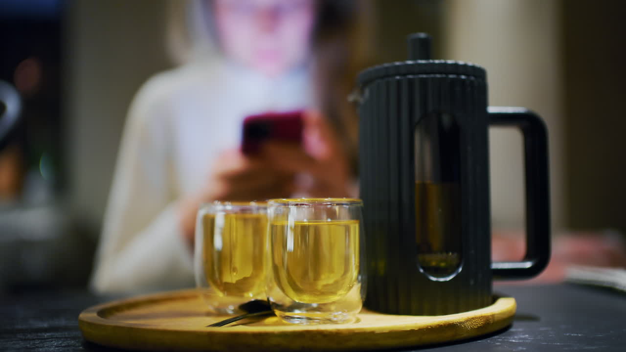 Due tazze di tè appena preparato su un vassoio, mentre una donna usa il suo smartphone sullo sfondo.